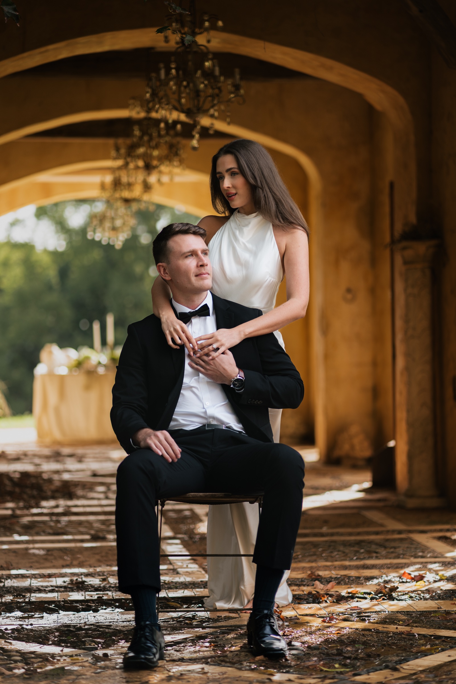 Bride standing behind the seated groom beneath a golden stone archway.