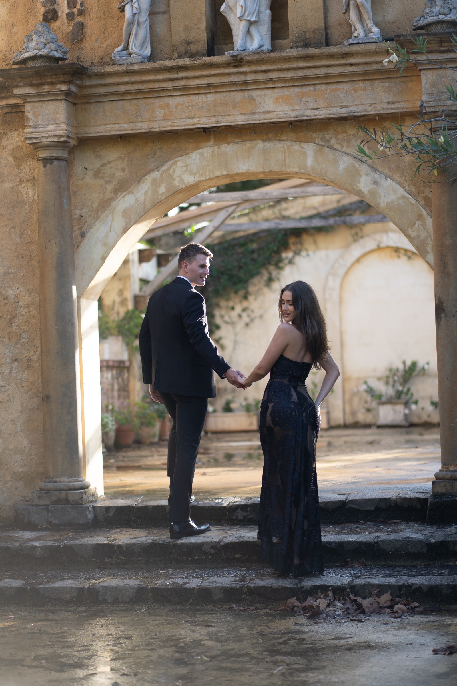Couple in black formalwear holding hands beneath a weathered stone arch.