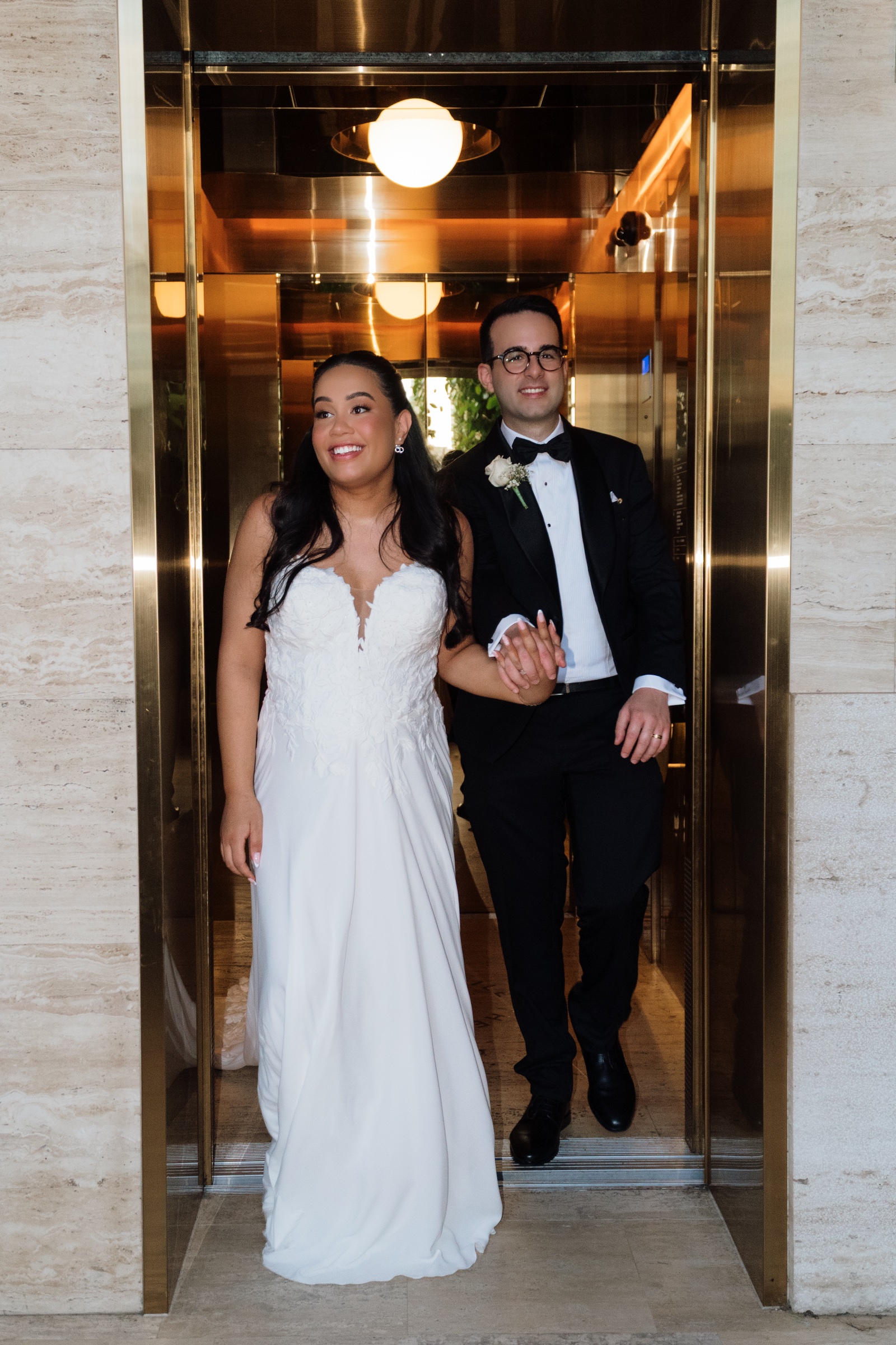 Bride and groom standing arm in arm in the doorway of a brass-toned lift.