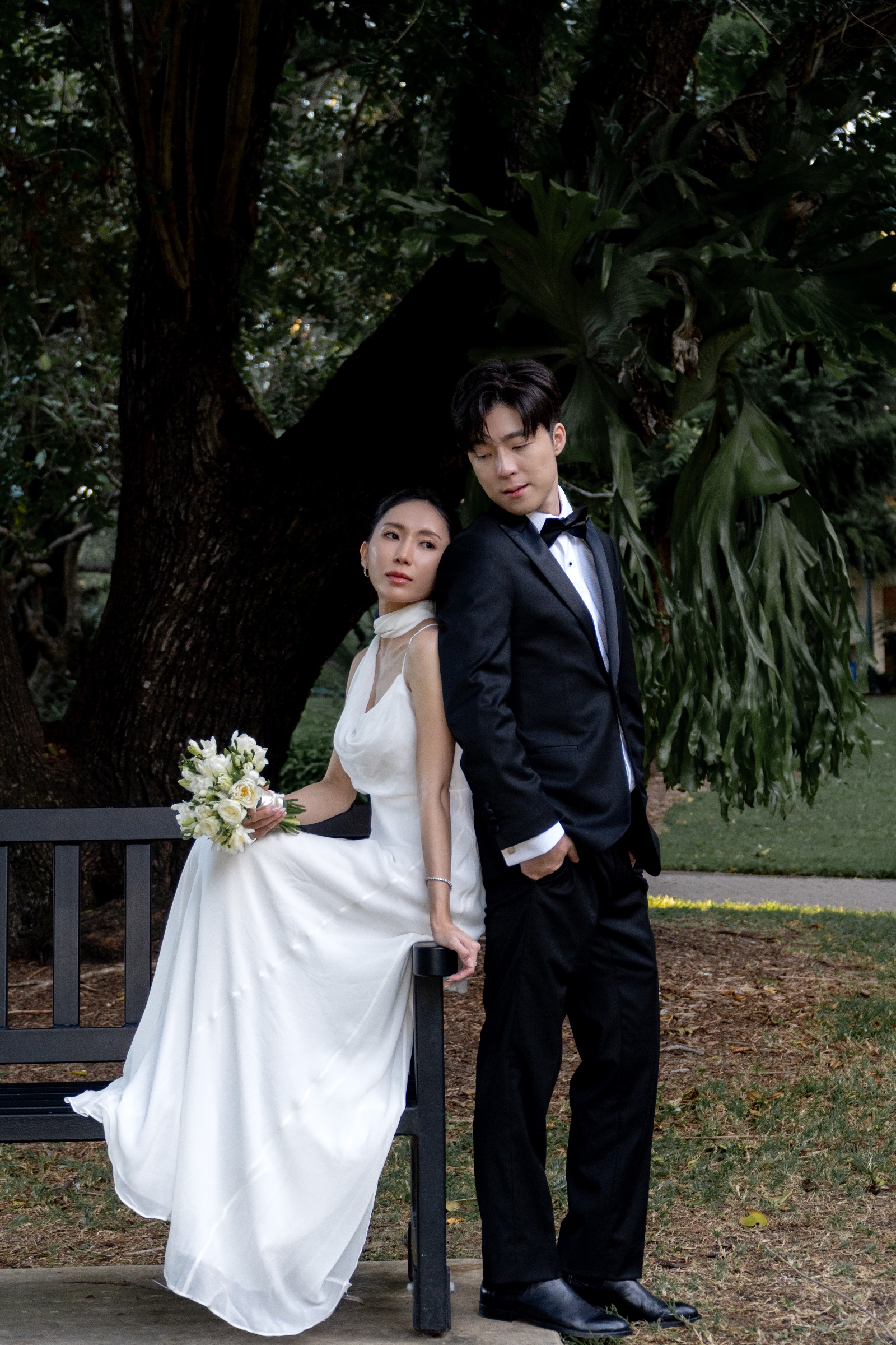 Couple in a dark tuxedo and white dress beside a garden bench, holding a bouquet.