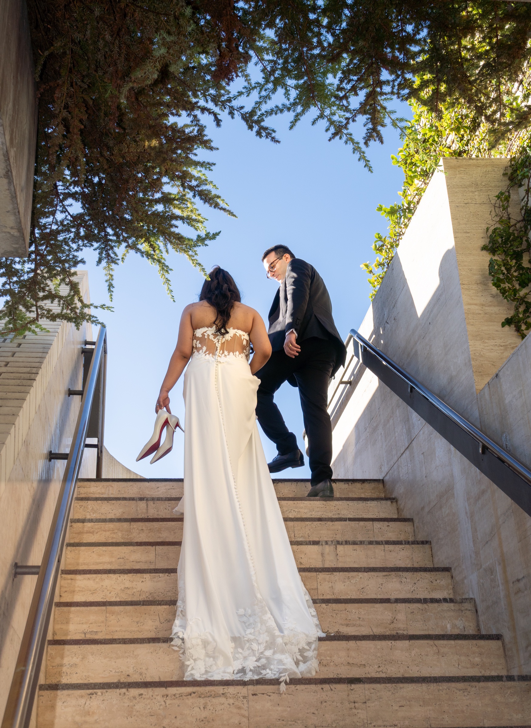 Bride walking up outdoor steps towards the groom beneath leafy branches.