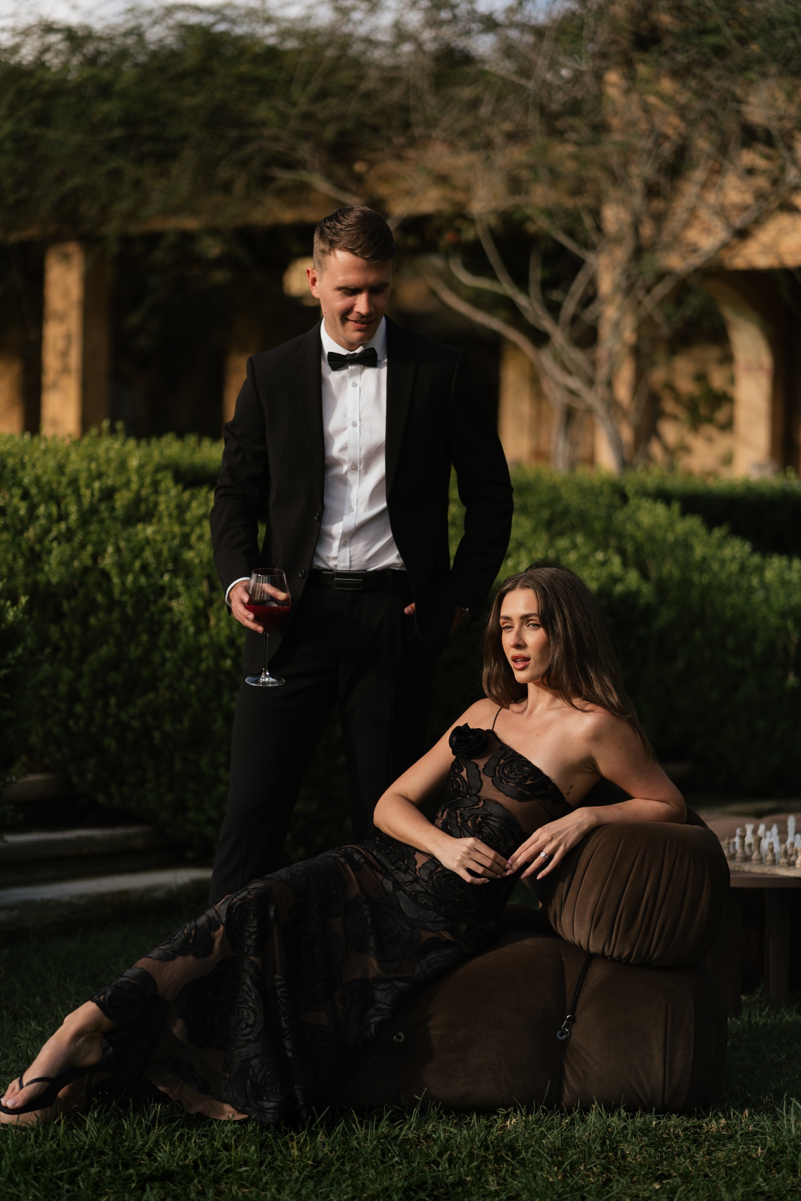 Couple in black evening wear posing beside a chair in a leafy garden.