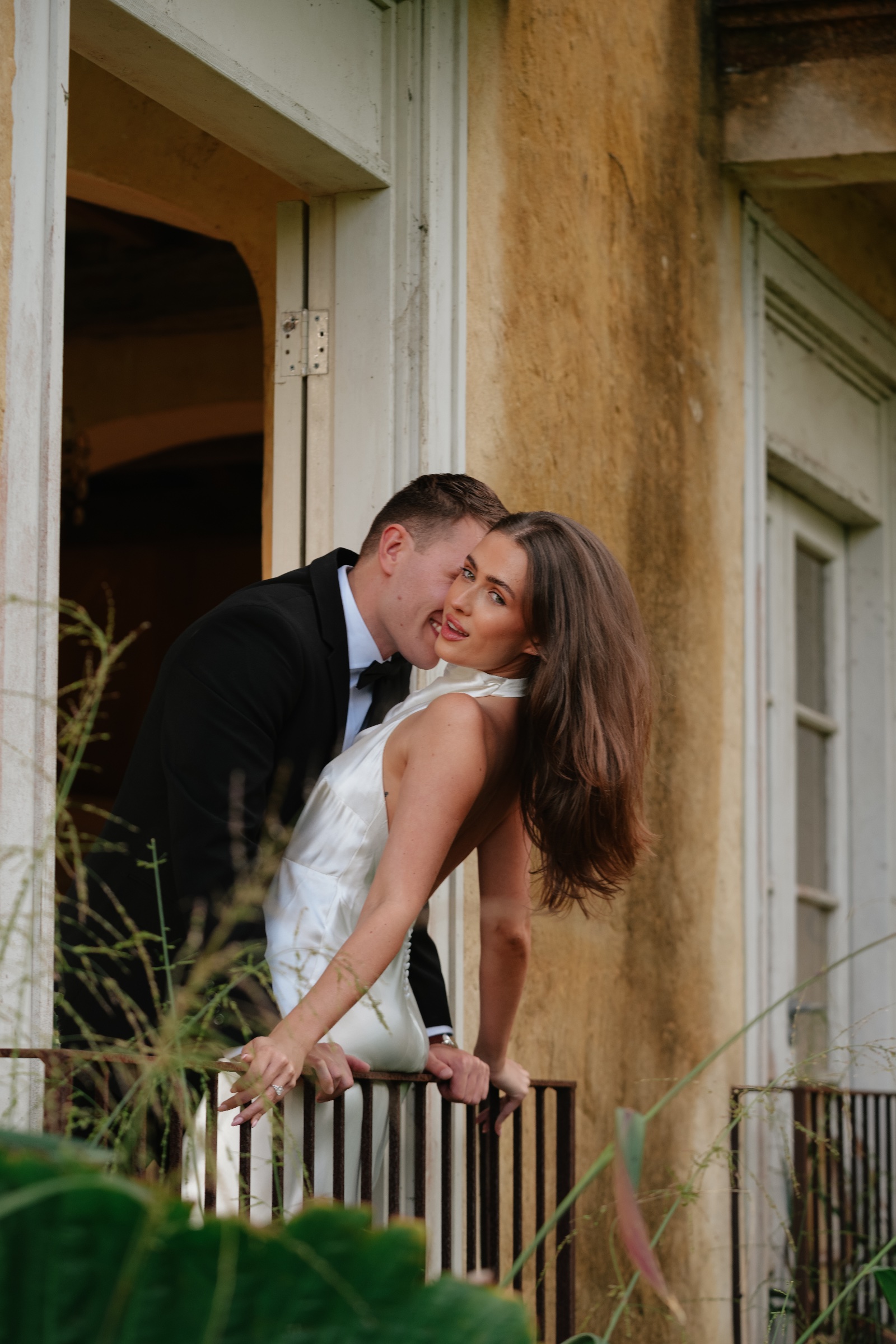 Couple leaning together beside a balcony railing on a warm-toned building.