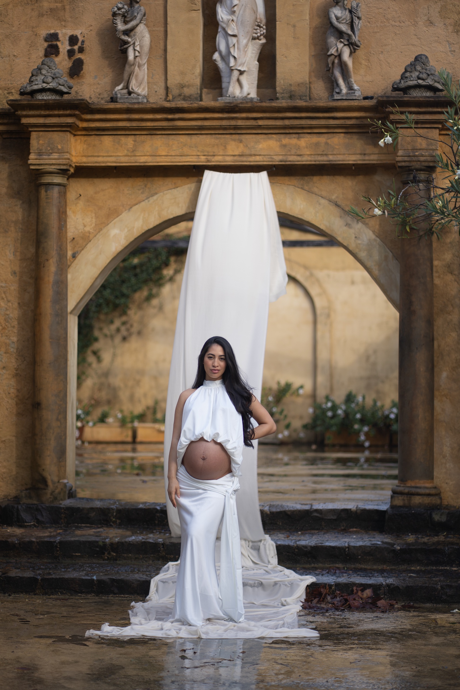 Maternity portrait in white fabric framed by a tall stone arch and hanging drapery.