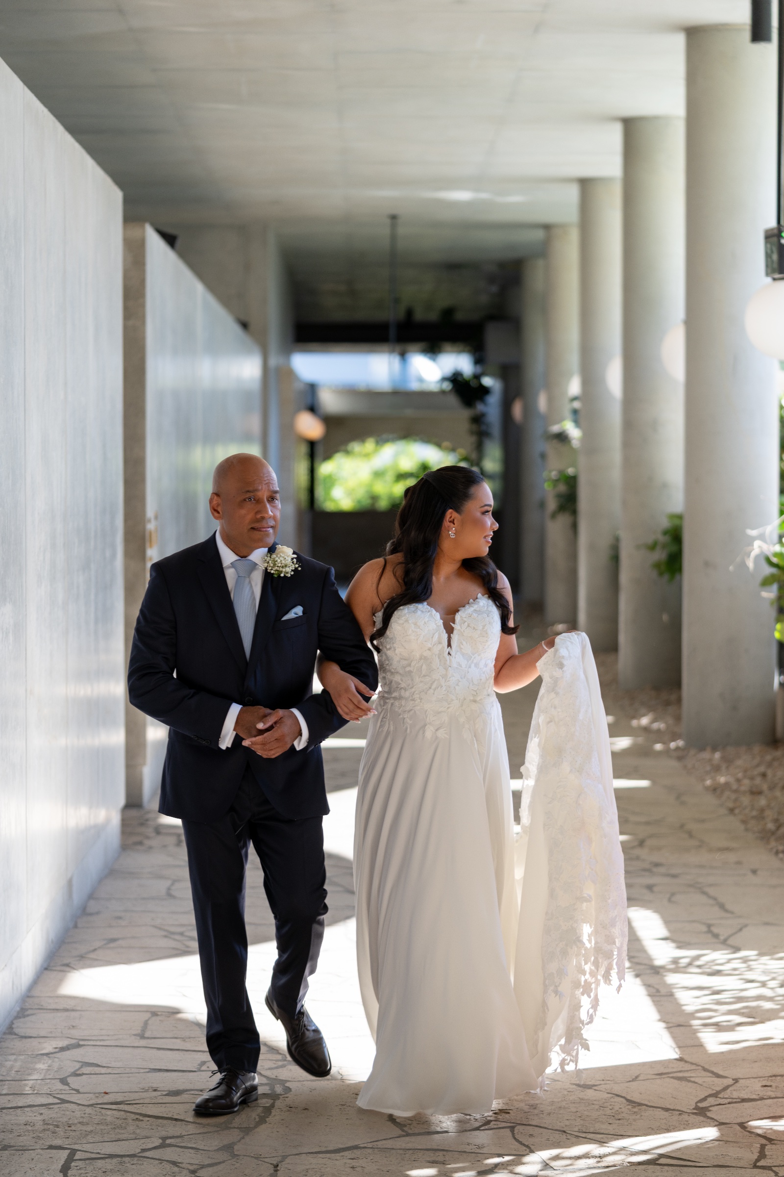 Bride and groom walking arm in arm through a sunlit covered passage.