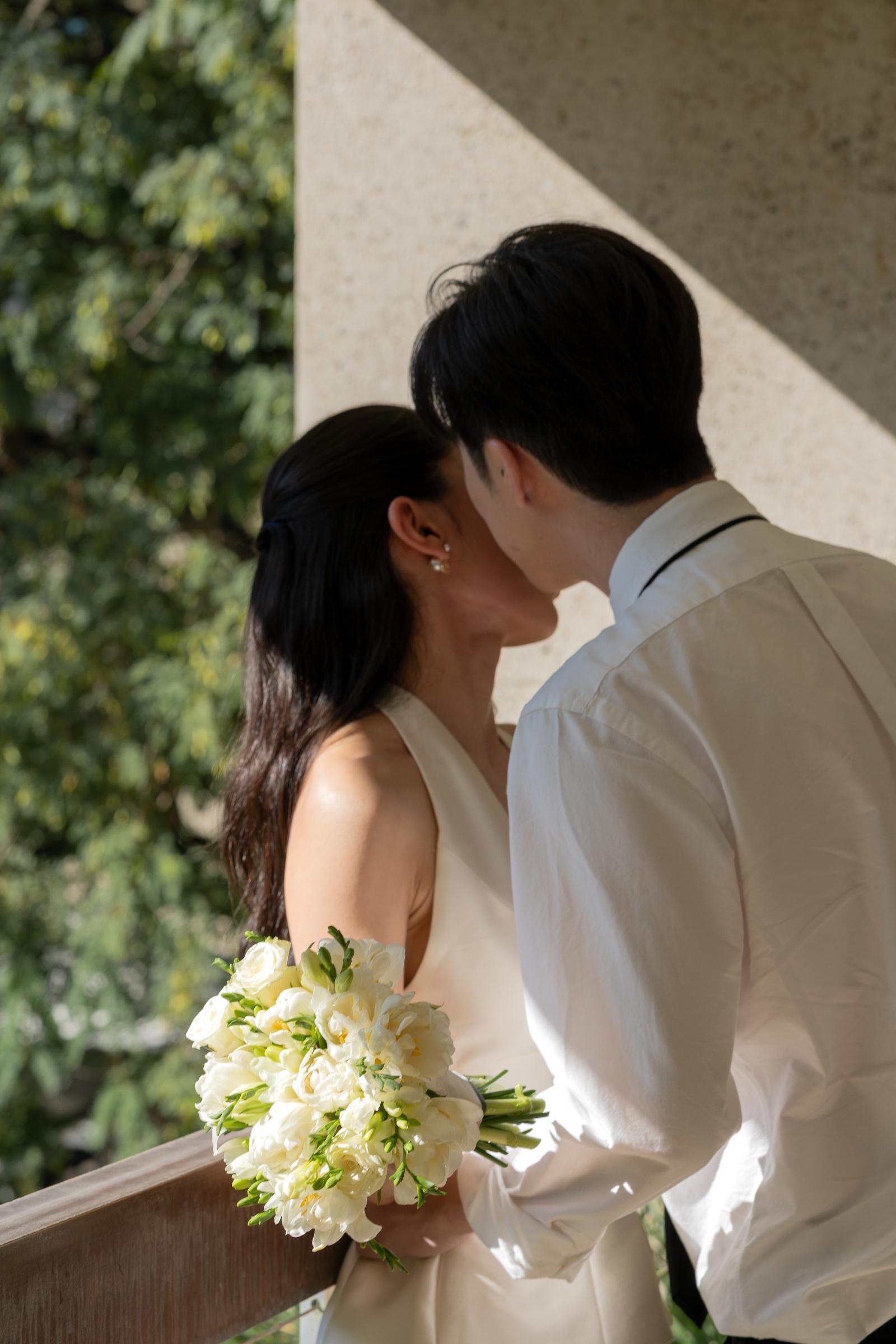 Couple kissing beside a stone column while holding a white and green bouquet.