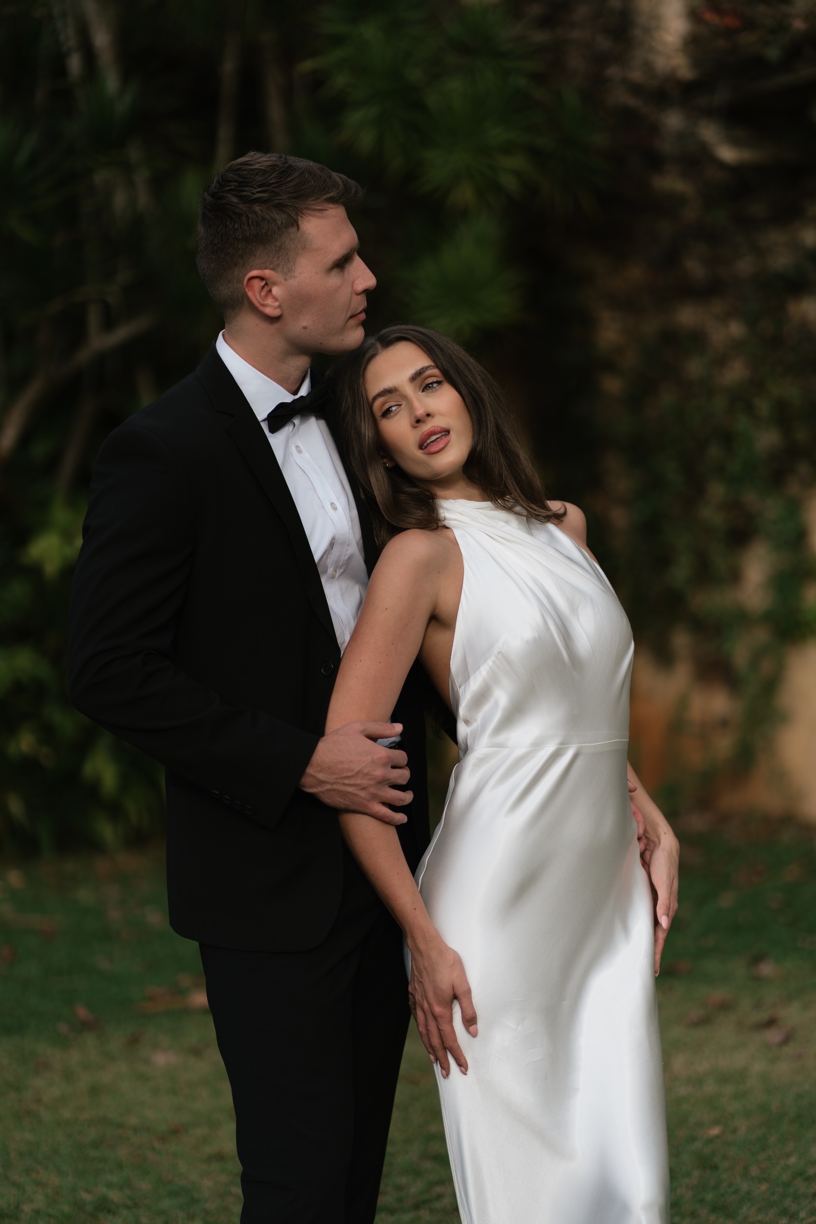 Couple in a black tuxedo and white halter-neck gown embracing in a leafy garden.