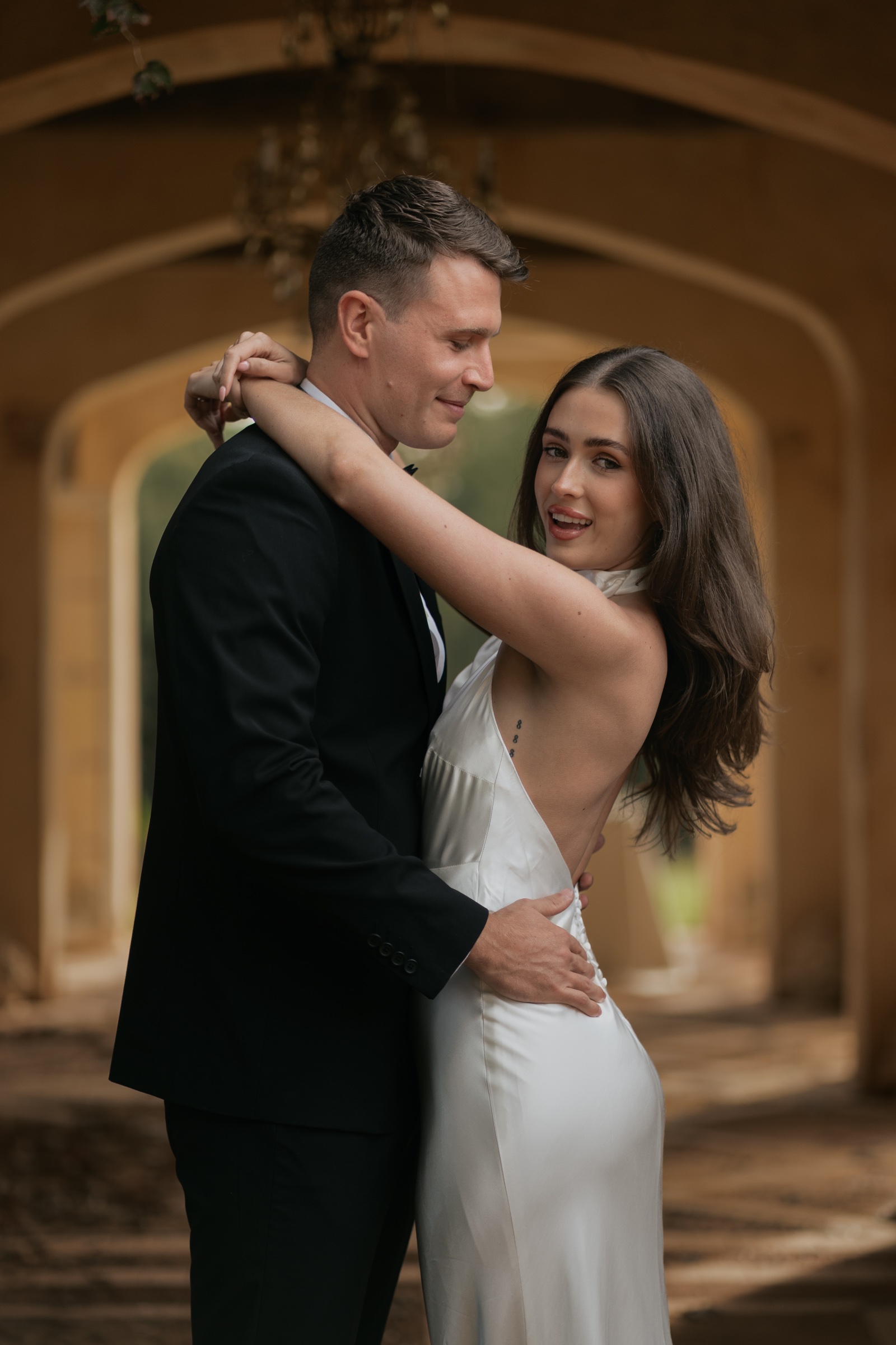 Bride and groom embracing beneath an arch, with the bride looking towards the camera.