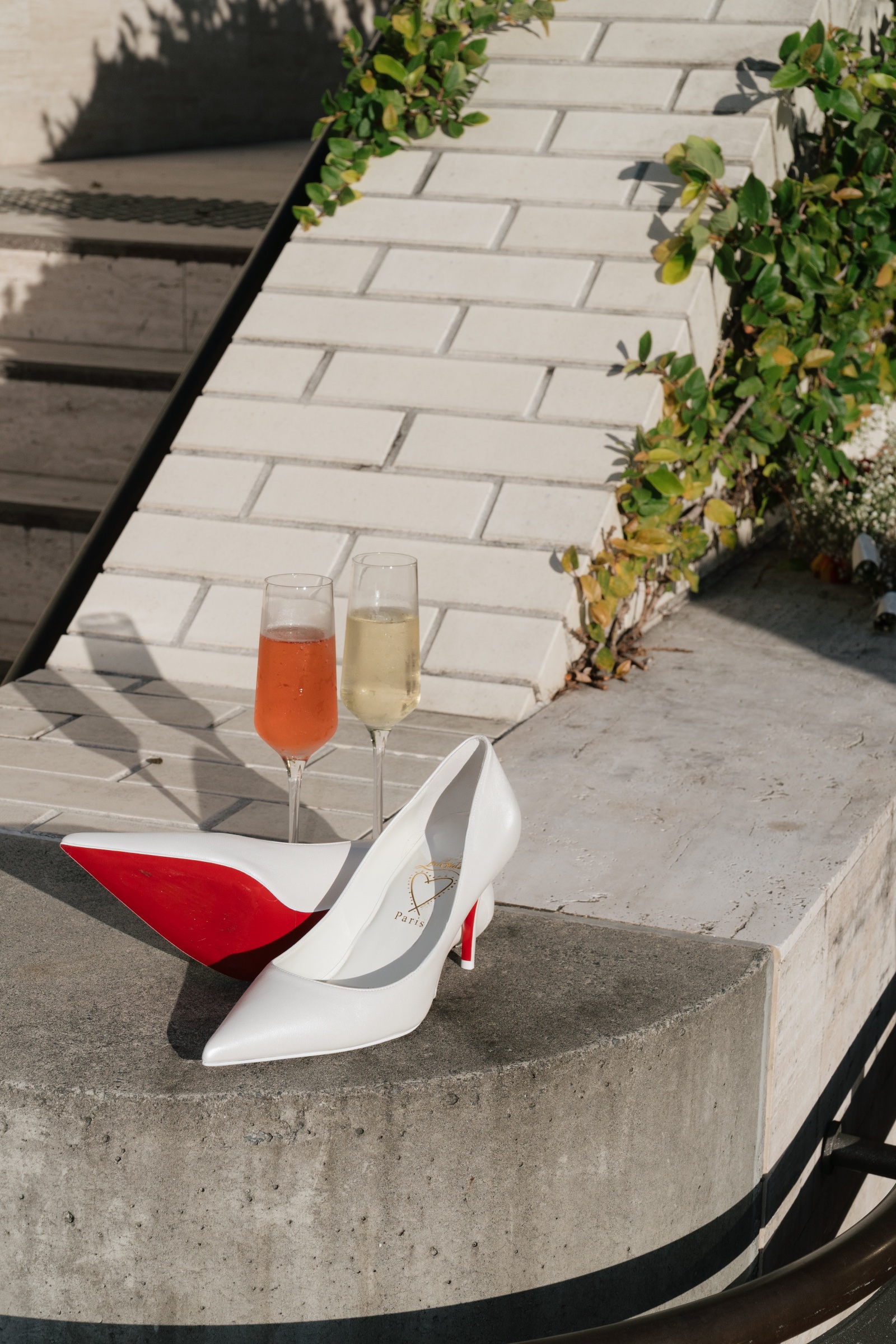 White bridal shoes with red soles beside two champagne glasses on a stone ledge.