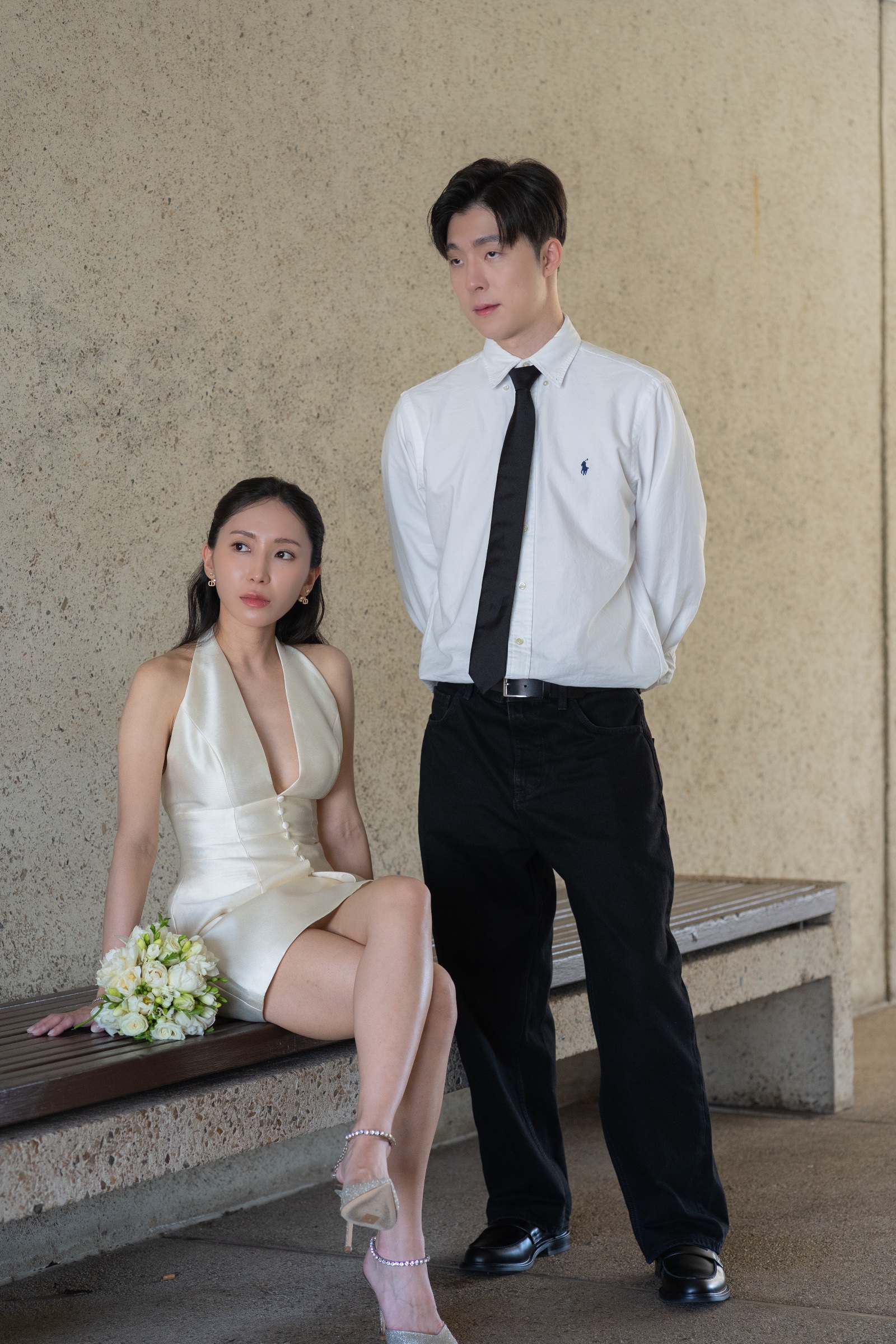 Couple posing beside a stone bench, with one partner seated and holding a bouquet.