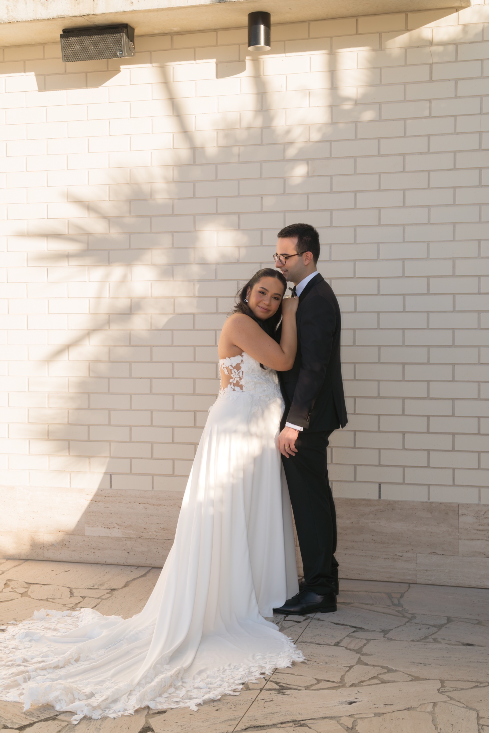 Bride and groom embracing beside a pale brick wall in warm sunlight.