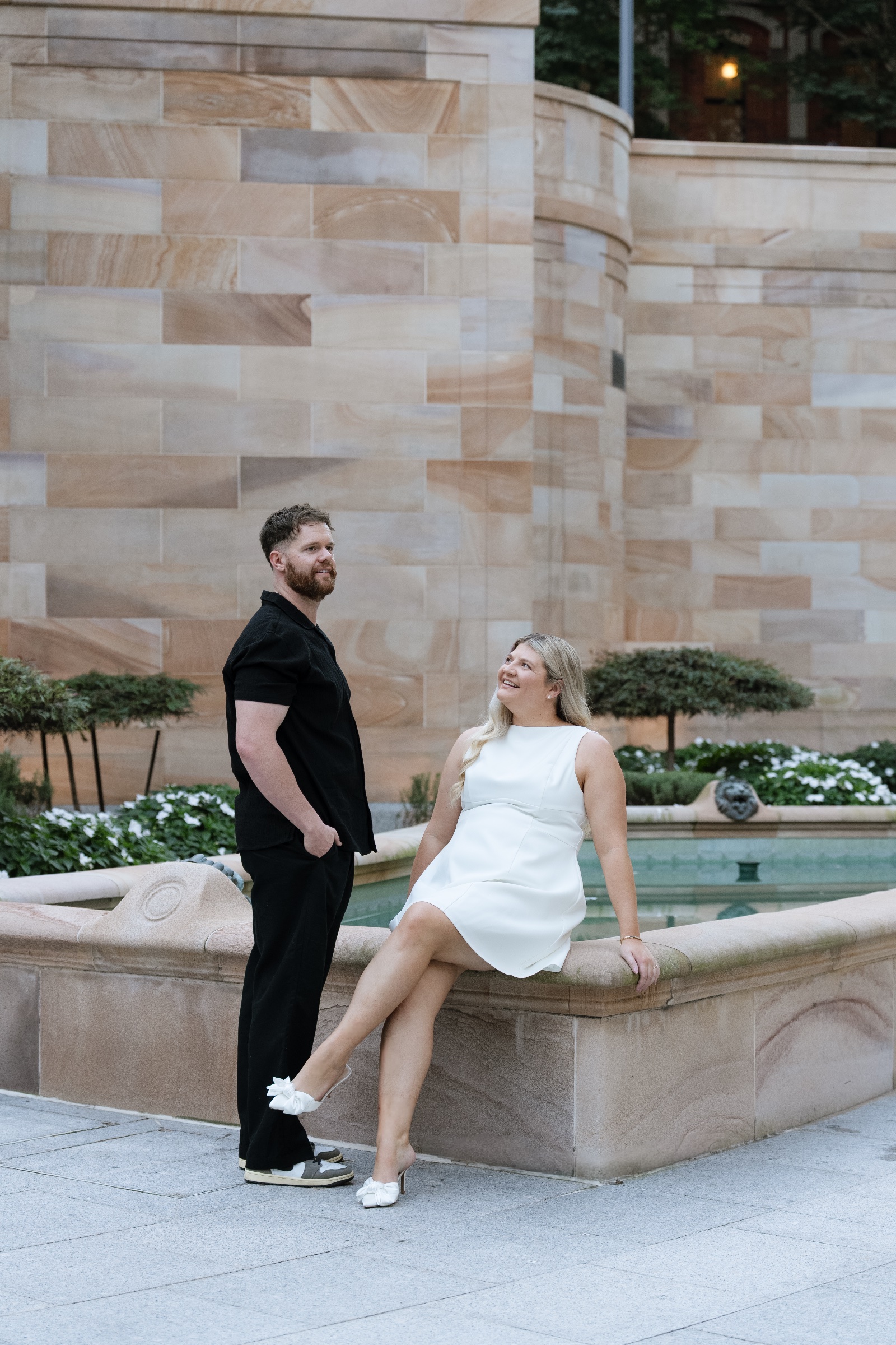 Couple beside a stone fountain, with one partner seated on the fountain edge.